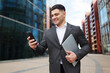 © muse studio - Businessman checks phone while smiling in urban area with modern buildings near midday