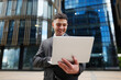 © muse studio - Businessman smiles while using laptop outside a modern building in the city center