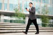 © muse studio - Young man walks confidently while looking at his smartphone outside a modern building