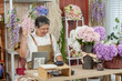 © Ekkasit A Siam - Asian senior female florist celebrating success while using digital tablet in flower shop, raising fist joyfully, surrounded by fresh hydrangeas, proud small businesswoman after retirement