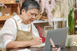 © Ekkasit A Siam - Asian senior florist writing in notebook while working on laptop at flower shop table filled with documents coffee and digital tools for managing small business after retirement