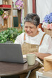 © Ekkasit A Siam - asian senior florist working at flower shop looking at laptop and smiling holding cup enjoying coffee break while checking business updates surrounded by plants tools and floral products
