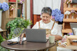 © Ekkasit A Siam - Smiling asian senior florist woman managing flower shop business using laptop while enjoying peaceful moment at cozy workspace surrounded by flowers and positive online sales results