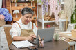 © Ekkasit A Siam - Elderly asian florist checking sales data on laptop smiling with excitement showing successful performance of online flower business and positive customer response