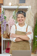 © Ekkasit A Siam - Senior Asian female florist smiling confidently with arms crossed in front of laptop inside flower shop with handmade floral arrangements and decorative flowers in soft tones