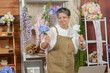 © Ekkasit A Siam - Smiling asian senior female florist holding artificial flowers dancing in flower shop workplace filled with colorful arrangements enjoying small business success and joyful online interaction