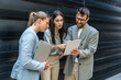 © Srdjan - Three AI innovation experts meeting outside office building, two businesswomen and businessman discussing strategy with tablet and laptop, smiling during modern tech consulting networking