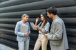 © Srdjan - Three AI innovation experts meeting outside office building, two businesswomen and businessman discussing strategy with tablet and laptop, smiling during modern tech consulting networking