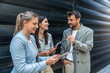© Srdjan - Three AI innovation experts meeting outside office building, two businesswomen and businessman discussing strategy with tablet and laptop, smiling during modern tech consulting networking
