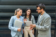 © Srdjan - Three AI innovation experts meeting outside office building, two businesswomen and businessman discussing strategy with tablet and laptop, smiling during modern tech consulting networking