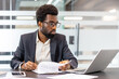 © Liubomir - Concentrated professional man reviewing documents and working on a laptop at a modern office desk, managing business affairs and planning new strategies