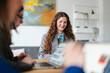 © Studio Marmellata - A young woman smiles while participating in a business meeting with colleagues, discussing projects on laptops in a modern office setting.