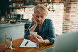 © Davor - Senior woman looking at smartphone with bills in home kitchen
