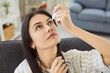 © Studio Romantic - Portrait of young woman sitting on sofa in living room, tilting head back and applying eye drops. Female using medication to relieve eyes dryness, irritation or inflammation and maintain eye health.
