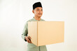 © Wayan - An Indonesian Muslim man in a green traditional shirt and black cap holds a brown cardboard box with a happy smiling expression against a white background. Quarter Side View