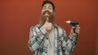 © Krakenimages.com - Man holding a cordless drill gives thumbs up in studio with a vibrant orange backdrop and wearing a plaid shirt; confidence.