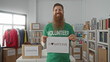 © Krakenimages.com - Bearded man in green volunteer shirt holds paper donation sign amid stacked boxes and clothing racks in building; compassion.
