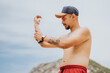 © qunica.com - A shirtless man in a dark cap and red shorts applies sunscreen on his arm at a rocky outdoor beach. Relaxed, sunny mood with a fitness-influenced, casual vibe.