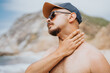 © qunica.com - A shirtless man on a sunny beach wears a cap and sunglasses, touching his neck in a thoughtful moment. The rocky shore and blue sky create a calm outdoor lifestyle vibe.