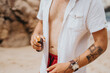© qunica.com - A man stands on the beach with an open white shirt and red shorts, holding a bottle of sunscreen. His tattooed arm and relaxed, casual pose convey summer skincare and outdoor leisure.
