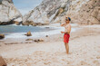 © qunica.com - A man in red swim shorts and a cap stands on a sandy beach, looking toward the ocean with a relaxed pose. Backpack rests on the sand as waves roll in.