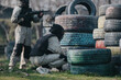 © qunica.com - Two players in camouflage and protective gear crouch behind stacked tires on a paintball field. A teammate aims a marker while others scout, capturing a tactical moment in the game.