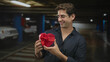 © Krakenimages.com - Man holding red heart gift box with both hands in a parking building, smiling while presenting the gift; romantic affection.