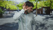 © Krakenimages.com - Hispanic man in white shirt cups ear on a sunlit city street lined with green trees and parked cars; curiosity.