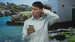 © Krakenimages.com - Young hispanic man scratches head holding smartphone beside a white building by the turquoise seaside; confusion.