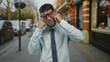 © Krakenimages.com - Hispanic man wearing round glasses and a blue shirt pressing cheeks with fists on city street in front of a brick building; toothache.