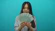 © Krakenimages.com - Young chinese woman holding banknotes with a curious expression against an isolated blue background, showcasing a fan of yuan money in her hands.