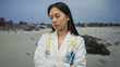© Krakenimages.com - Asian woman doctor in uniform with stethoscope stands on a scenic seaside beach, showcasing a blend of professional and serene outdoor environment.