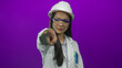 © Krakenimages.com - Young chinese woman in a hardhat and lab coat points forward against an isolated purple wall background, emphasizing her role in a professional or scientific setting.