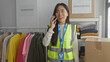 © Krakenimages.com - Young woman in reflective vest talks on phone in an indoor charity center, surrounded by donations of clothes and boxes, signifying volunteer work.