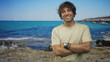 © Krakenimages.com - Man with crossed arms wearing watch and bracelets standing on a rocky beach facing camera with a slight smile and closed eyes; relaxed confidence.