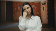 © Krakenimages.com - Hispanic woman standing thoughtfully indoors at a construction site, wearing a white shirt with a backdrop of unfinished brick walls and soft lighting, evoking a sense of contemplation.