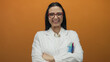 © Krakenimages.com - Young hispanic woman scientist in white uniform smiling confidently against vibrant orange wall background wearing glasses and crossing arms displaying professionalism.