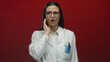 © Krakenimages.com - Hispanic woman scientist in white uniform with toothache expression stands against a red wall, indicating dental pain, discomfort, and medical concern.
