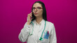 © Krakenimages.com - Hispanic female doctor in uniform with stethoscope and smartphone against a vibrant pink wall, conveying medical professionalism and communication.