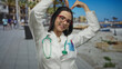 © Krakenimages.com - Young hispanic woman doctor in uniform making heart gesture with hands standing on seaside promenade with stethoscope outdoors showing positive emotion near the beach.