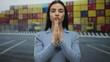 © Krakenimages.com - Woman praying outdoors at a port with colorful shipping containers, expressing hope and contemplation, wearing blue shirt, hispanic, young, under natural light in an industrial area.
