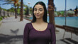 © Krakenimages.com - Young woman with dark hair stands on a sunny promenade by the seaside, radiating confidence against a backdrop of palm trees and a serene sea.