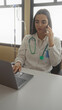 © Krakenimages.com - Young hispanic woman doctor in uniform with stethoscope using laptop and phone indoors in hospital room