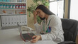 © Krakenimages.com - Woman doctor in hospital room removes glasses appearing stressed in front of laptop, with stethoscope visible, conveying work fatigue and healthcare pressures.