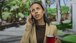 © Krakenimages.com - Woman talking on phone while holding red cup in sunny park surrounded by green nature, appearing thoughtful and intense in casual outdoor setting.