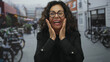 © Krakenimages.com - Hispanic woman with curly hair and glasses expressing surprise on a bustling urban street, surrounded by blurred bicycles and buildings in the background.