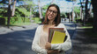 © Krakenimages.com - Woman wearing glasses holds stacked notebooks and folders against her chest while smiling on a shaded tree lined street with sunlit pavement; confidence study.