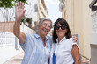 © luciano - Two happy senior women smiling and waving at the camera while walking together on a sunny residential street. The elderly friends embrace warmly, expressing joy, friendship, and an active lifestyle
