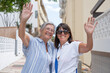 © luciano - Two happy senior women smiling and waving at the camera while walking together on a sunny residential street. The elderly friends embrace warmly, expressing joy, friendship, and an active lifestyle