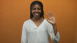 © Krakenimages.com - African american woman smiling and waving hand in orange studio, wearing white blouse and braided hair, facing camera with nose ring; warmth welcome.